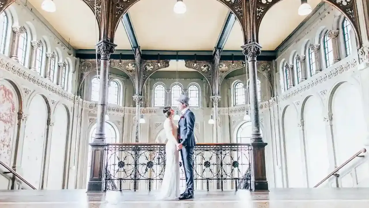 Ein Brautpaar im Stil der 20er Jahre steht sich in einem prachtvollen Gebäude mit Jugendstil-Architektur gegenüber.