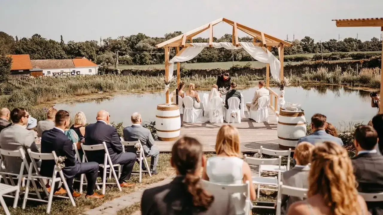 Eine romantische **Hochzeit im Freien** am See. Gäste sitzen auf Stühlen, das Brautpaar steht unter einem Holzdach.