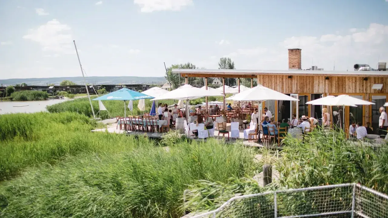 Gäste genießen eine festliche Hochzeit am Wasser. Sonnenschirme spenden Schatten, Tische sind für das Festessen gedeckt.