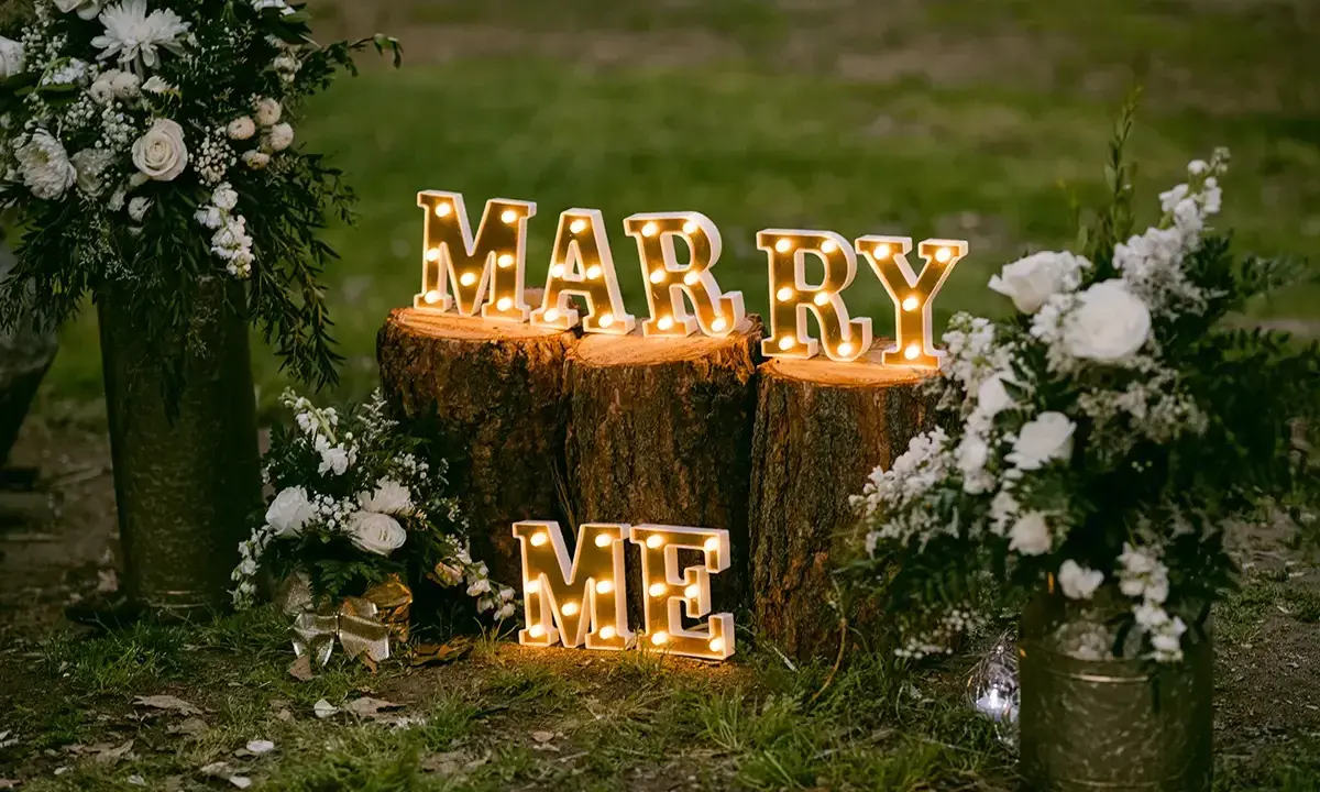 Leuchtende Buchstaben "MARRY ME" auf Baumstämmen, umgeben von Blumen. Perfekte heiratsantrag ideen für einen romantischen Moment.