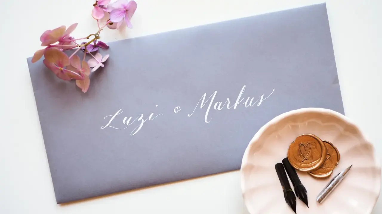 Grauer Umschlag mit weißer Kalligrafie "Luzi & Markus". Daneben eine rosa Schale mit Siegelwachs, Stiften und Federn. Was schreibt man auf den Umschlag zur Hochzeit?