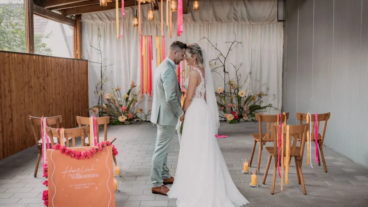 Brautpaar im boho style wedding, umgeben von Blumen und Bändern. Ein Willkommensschild steht im Vordergrund.