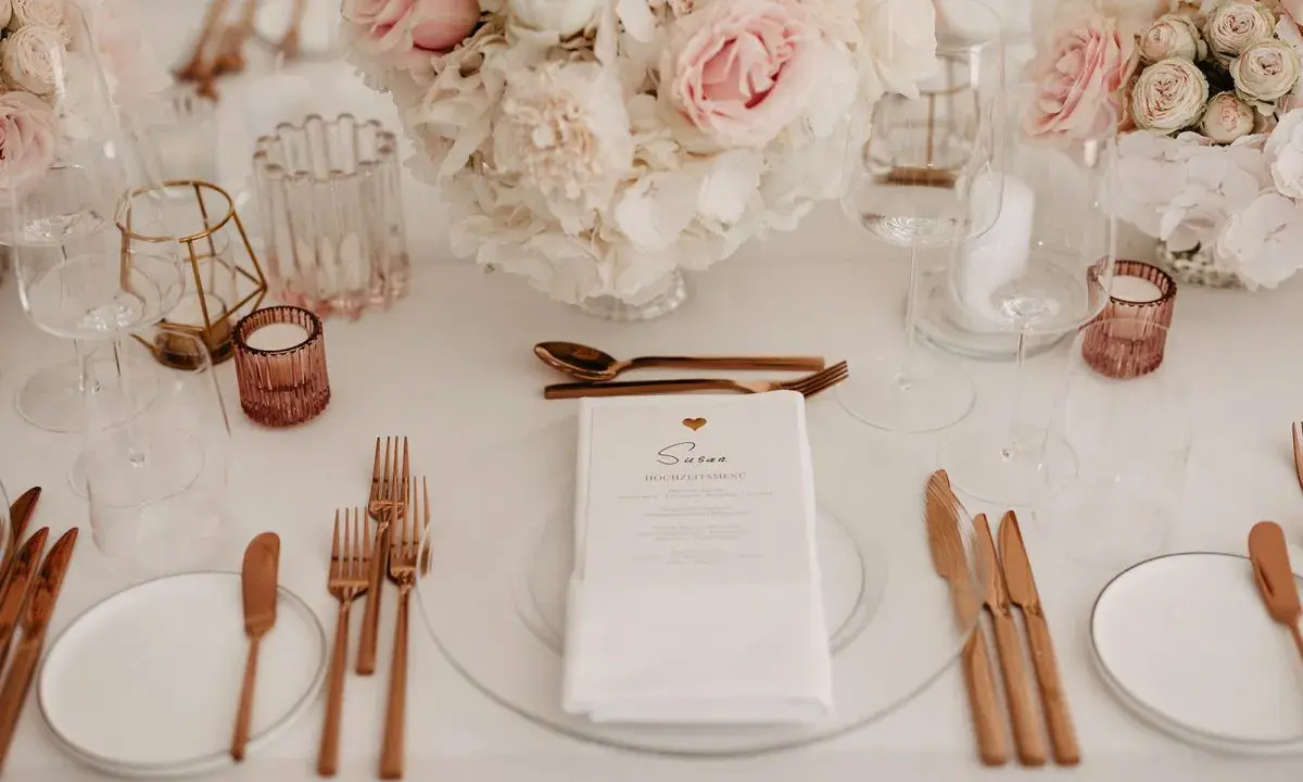 Ein festlich gedeckter Tisch mit Blumen, Gläsern und roségoldenem Besteck. Ein Hochzeitsmenü für Susan, perfekt arrangiert vom wedding planer.