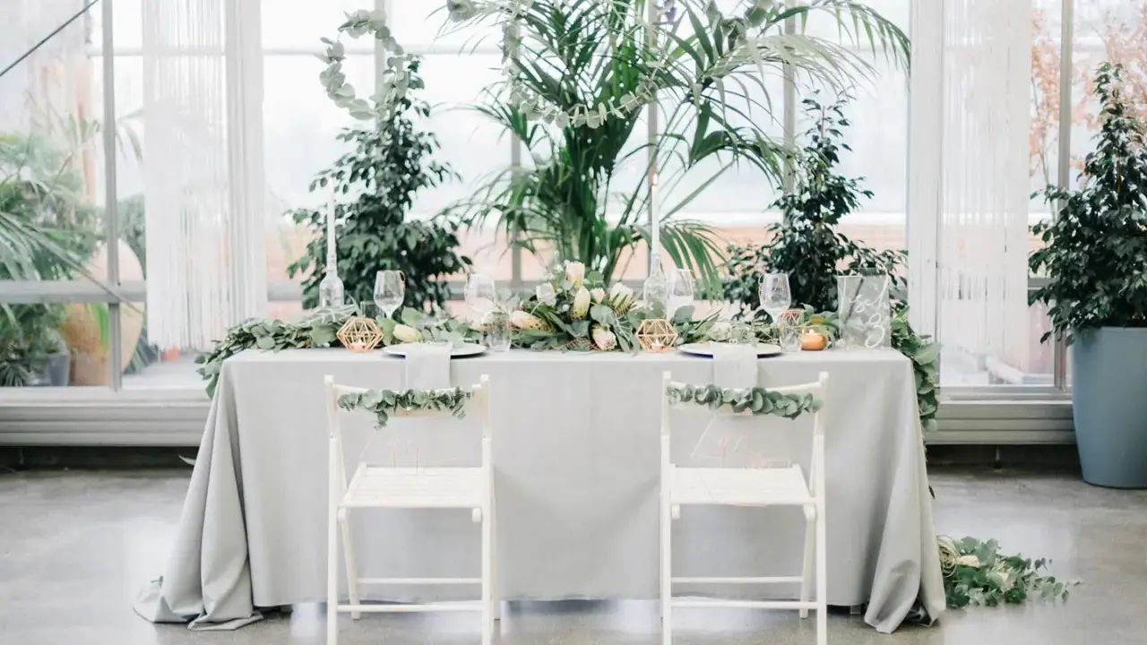 Ein festlich gedeckter Tisch für eine greenery hochzeit, umgeben von üppigem Grün. Zwei weiße Stühle mit Eukalyptus-Girlanden stehen bereit.