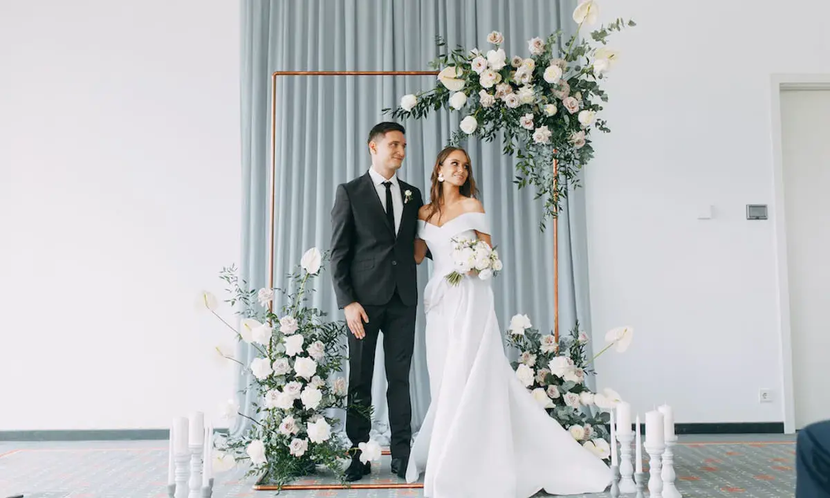 Ein Brautpaar steht vor einem floralen Bogen, bereit, wie heiratet man. Sie strahlen Glück aus.