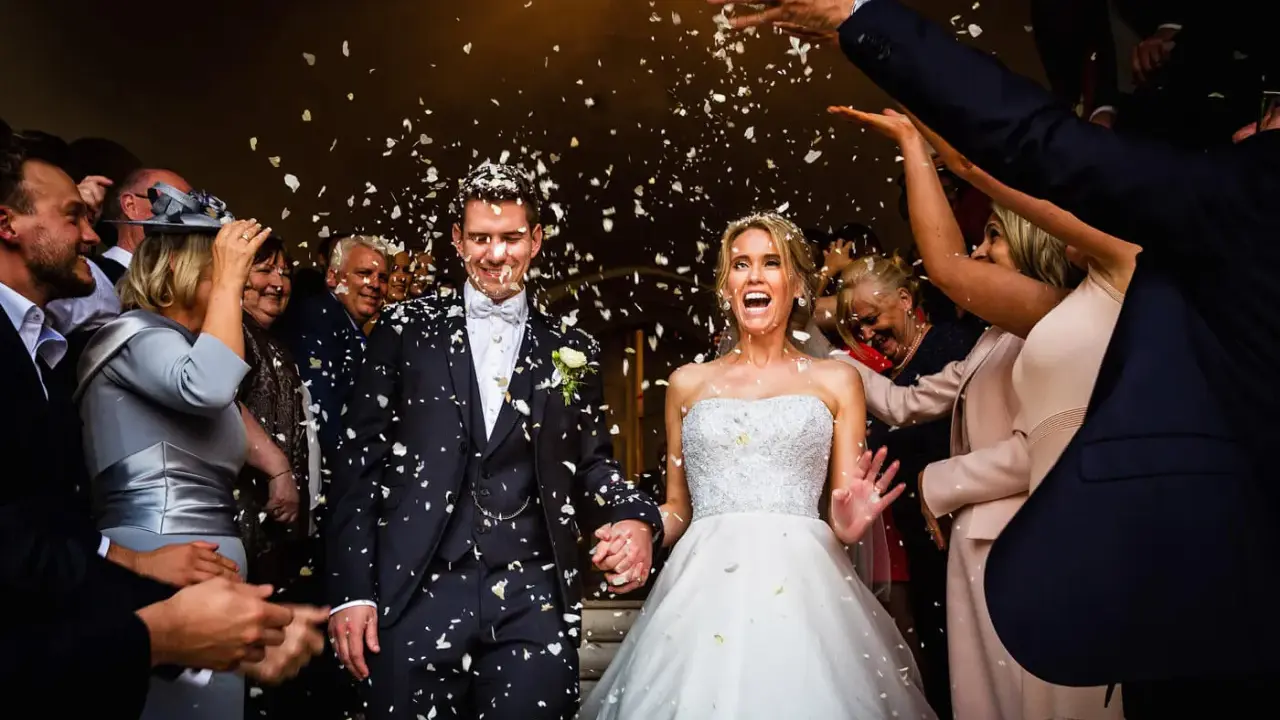 Glückliches Brautpaar bei einer englischen Hochzeit, umgeben von Blütenblättern und jubelnden Gästen.