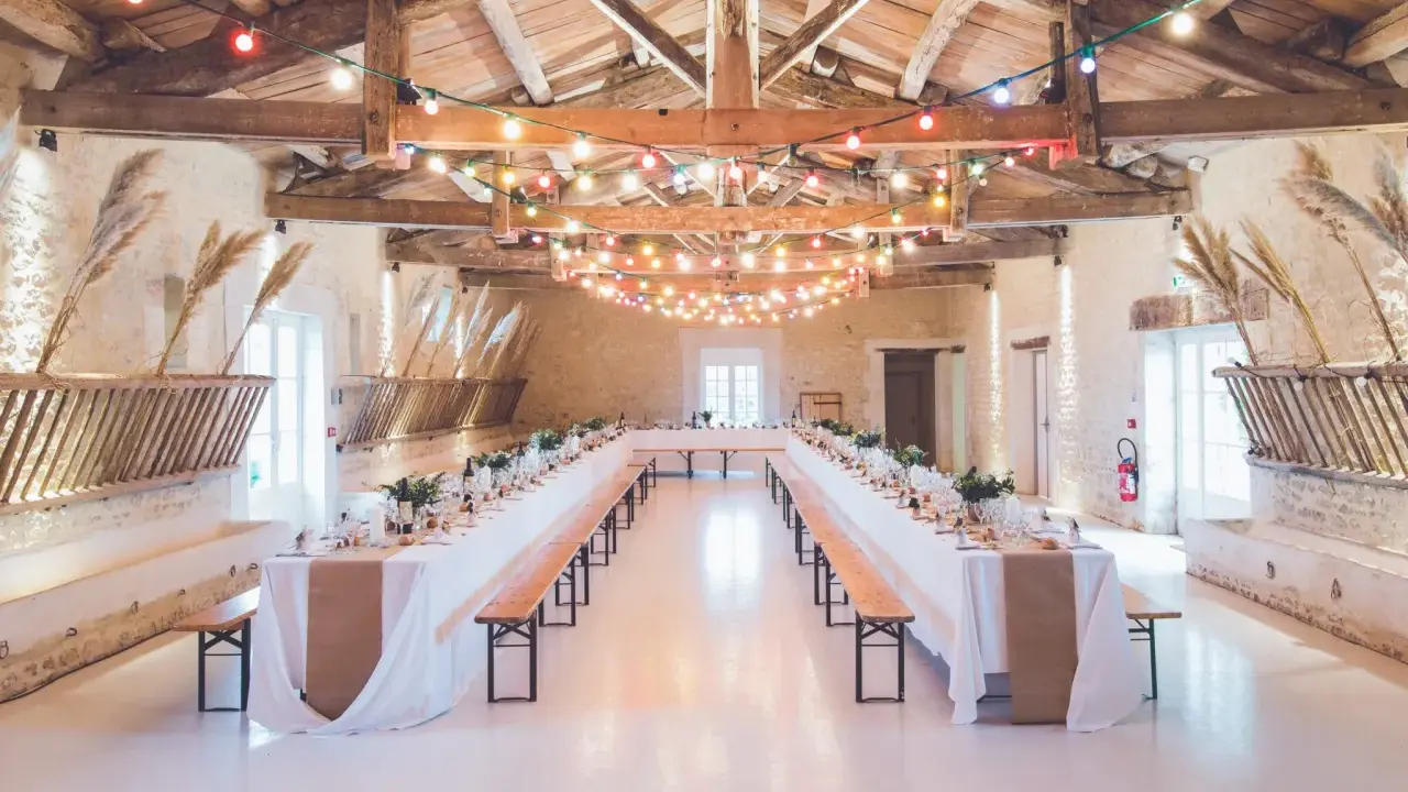 Festlich geschmückte alte Scheune für eine Hochzeit, mit langen Tafeln, Bänken und bunten Lichterketten unter Holzbalken.