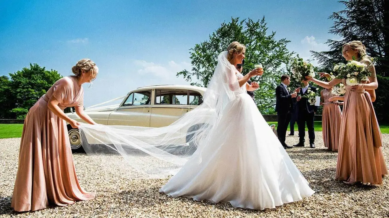 Brautjungfern helfen der Braut mit ihrem Schleier vor einer Hochzeitslimousine.