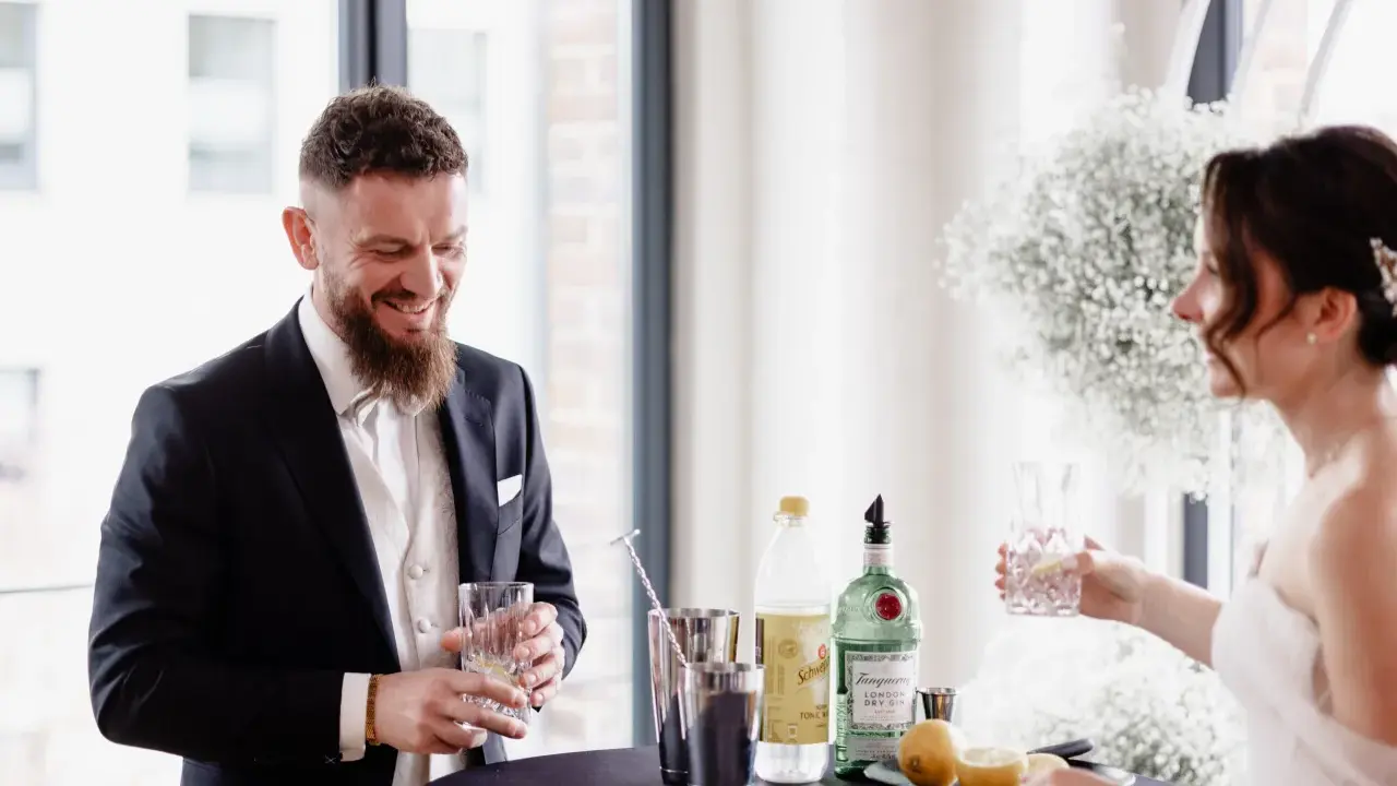 Ein lachender Bräutigam mit einem Drink in der Hand, während die Braut ihm ein Glas reicht. Ein Moment der Freude vor ihrer rituale freie trauung.
