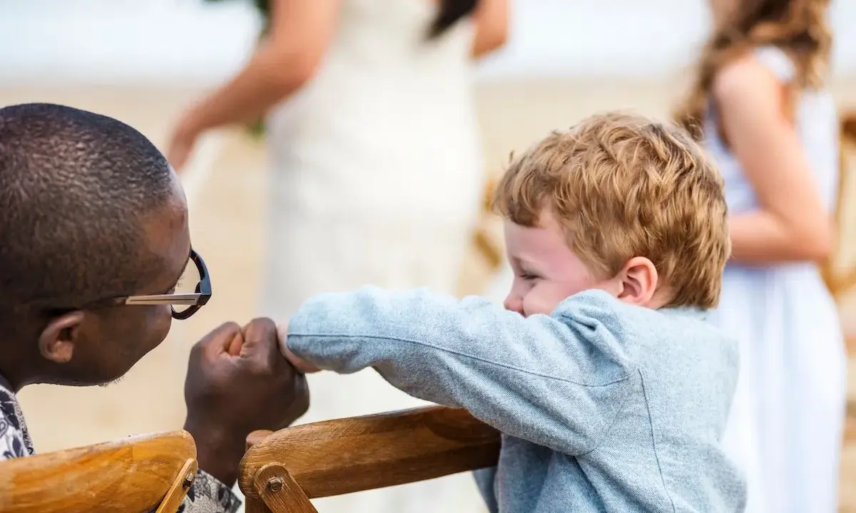 Ein Mann mit Brille hält die Hand eines Jungen. Im Hintergrund heiratet mein Sohn.