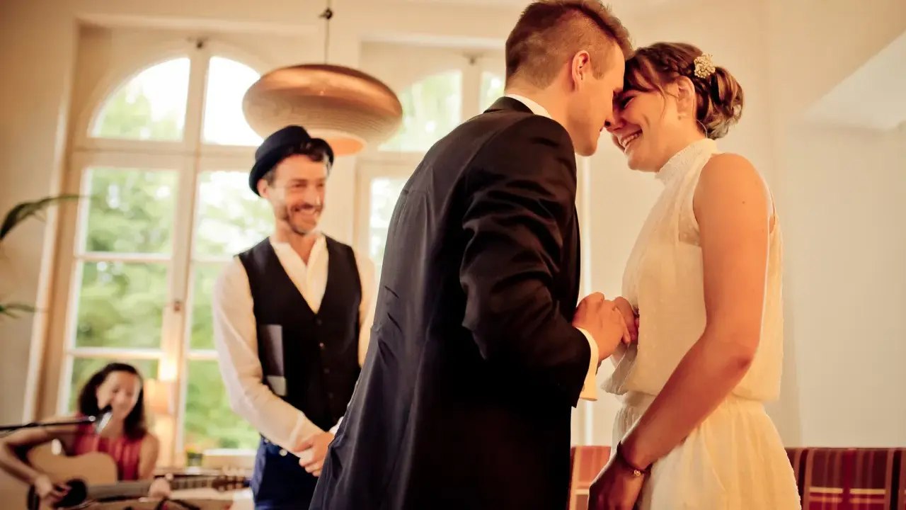 Ein Brautpaar teilt einen innigen Moment, während ein Mann mit Hut und eine Frau mit Gitarre im Hintergrund stehen. Die emotionale Rede beim Standesamt wird durch ihre Freude spürbar.