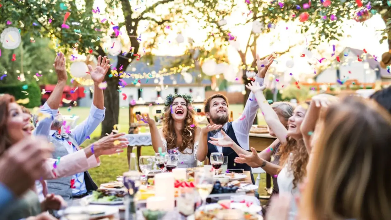 Glückliche Gäste werfen Konfetti bei einer fröhlichen Garten Hochzeit. Das Brautpaar lacht ausgelassen.