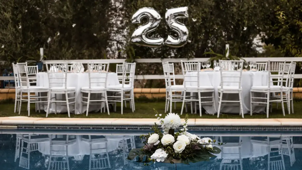Festlich gedeckte Tische mit weißen Stühlen und Luftballons "25" über ihnen – perfekt für eine Rede zur Silberhochzeit am Pool.