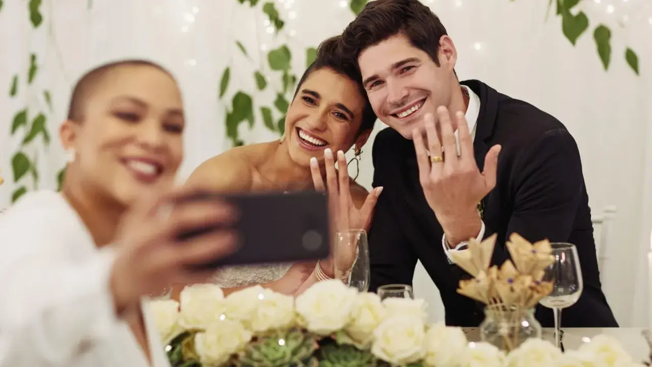 Brautpaar zeigt Ringe für ein Hochzeitsfoto, während eine Freundin ein Selfie macht.