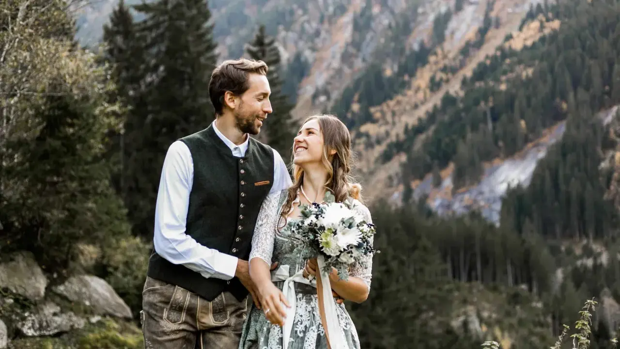 Ein glückliches Brautpaar feiert seine Hochzeit auf der Alm. Sie tragen traditionelle Tracht vor einer Bergkulisse.