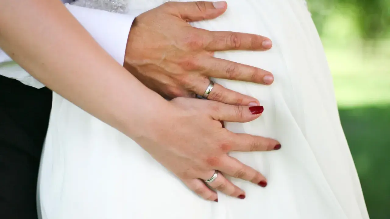 Hände auf dem Bauch einer schwangeren Braut im weißen Kleid. Ein Paar heiratet voller Vorfreude.