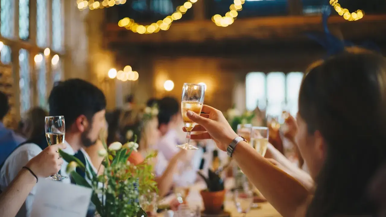 Gäste stoßen mit Sekt an, Lichterketten leuchten. Eine Einladung zur after wedding party.