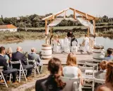 Eine romantische **Hochzeit im Freien** am See. Gäste sitzen auf Stühlen, das Brautpaar steht unter einem Holzdach.
