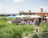 Gäste genießen eine festliche Hochzeit am Wasser. Sonnenschirme spenden Schatten, Tische sind für das Festessen gedeckt.