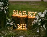 Leuchtende Buchstaben "MARRY ME" auf Baumstämmen, umgeben von Blumen. Perfekte heiratsantrag ideen für einen romantischen Moment.