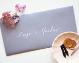 Grauer Umschlag mit weißer Kalligrafie "Luzi & Markus". Daneben eine rosa Schale mit Siegelwachs, Stiften und Federn. Was schreibt man auf den Umschlag zur Hochzeit?