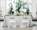 Ein festlich gedeckter Tisch für eine greenery hochzeit, umgeben von üppigem Grün. Zwei weiße Stühle mit Eukalyptus-Girlanden stehen bereit.