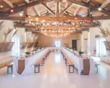 Festlich geschmückte alte Scheune für eine Hochzeit, mit langen Tafeln, Bänken und bunten Lichterketten unter Holzbalken.