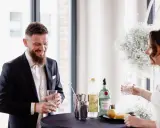 Ein lachender Bräutigam mit einem Drink in der Hand, während die Braut ihm ein Glas reicht. Ein Moment der Freude vor ihrer rituale freie trauung.