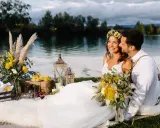 Glückliches Brautpaar genießt seine Hochzeit am See. Blumen, Laternen und ein Picknickkorb schaffen romantische Atmosphäre.