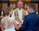 Ein glückliches Paar bei einer christlichen Hochzeit, umgeben von der feierlichen Atmosphäre einer Kirche. Der Bräutigam im blauen Anzug und die Braut im Spitzenkleid blicken sich an, während der Pfarrer sie segnet.
