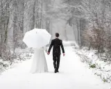 Ein Brautpaar geht Hand in Hand unter einem weißen Schirm durch einen verschneiten Wald. Vielleicht ist dies der beste Monat zum Heiraten für alle, die eine romantische Winterhochzeit wünschen.