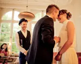 Ein Brautpaar teilt einen innigen Moment, während ein Mann mit Hut und eine Frau mit Gitarre im Hintergrund stehen. Die emotionale Rede beim Standesamt wird durch ihre Freude spürbar.