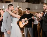 Glückliches Brautpaar umarmt sich, während ein Gitarrist spielt und Gäste applaudieren. Die Stimmung ist festlich, wie bei schönen Hochzeitsliedern auf Deutsch.