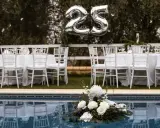 Festlich gedeckte Tische mit weißen Stühlen und Luftballons "25" über ihnen – perfekt für eine Rede zur Silberhochzeit am Pool.