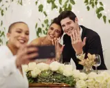 Brautpaar zeigt Ringe für ein Hochzeitsfoto, während eine Freundin ein Selfie macht.
