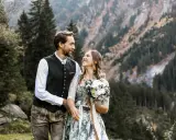 Ein glückliches Brautpaar feiert seine Hochzeit auf der Alm. Sie tragen traditionelle Tracht vor einer Bergkulisse.