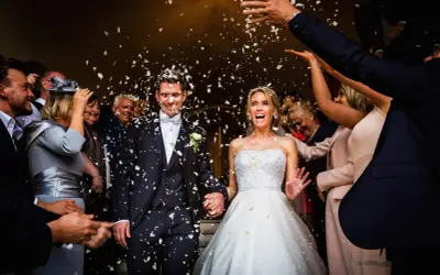 Glückliches Brautpaar bei einer englischen Hochzeit, umgeben von Blütenblättern und jubelnden Gästen.