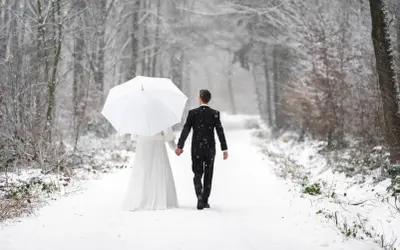 Ein Brautpaar geht Hand in Hand unter einem weißen Schirm durch einen verschneiten Wald. Vielleicht ist dies der beste Monat zum Heiraten für alle, die eine romantische Winterhochzeit wünschen.