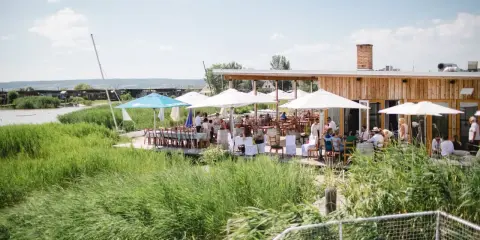 Gäste genießen eine festliche Hochzeit am Wasser. Sonnenschirme spenden Schatten, Tische sind für das Festessen gedeckt.