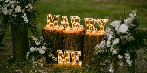 Leuchtende Buchstaben "MARRY ME" auf Baumstämmen, umgeben von Blumen. Perfekte heiratsantrag ideen für einen romantischen Moment.