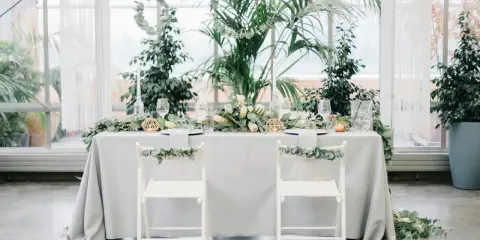 Ein festlich gedeckter Tisch für eine greenery hochzeit, umgeben von üppigem Grün. Zwei weiße Stühle mit Eukalyptus-Girlanden stehen bereit.