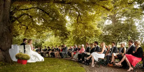 Hochzeit im Freien unter einem Baum. Gäste sitzen auf Bänken. Tolle Ideen zur Hochzeit für eine romantische Zeremonie.