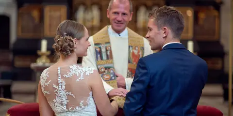 Ein glückliches Paar bei einer christlichen Hochzeit, umgeben von der feierlichen Atmosphäre einer Kirche. Der Bräutigam im blauen Anzug und die Braut im Spitzenkleid blicken sich an, während der Pfarrer sie segnet.