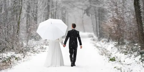 Ein Brautpaar geht Hand in Hand unter einem weißen Schirm durch einen verschneiten Wald. Vielleicht ist dies der beste Monat zum Heiraten für alle, die eine romantische Winterhochzeit wünschen.