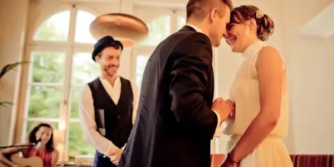 Ein Brautpaar teilt einen innigen Moment, während ein Mann mit Hut und eine Frau mit Gitarre im Hintergrund stehen. Die emotionale Rede beim Standesamt wird durch ihre Freude spürbar.