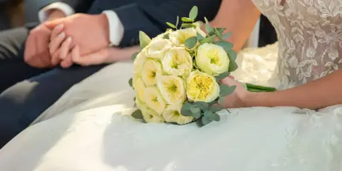 Brautpaar hält Händchen, Brautstrauß mit gelben Rosen. Eine Hochzeit Versicherung gibt Sicherheit für den großen Tag.