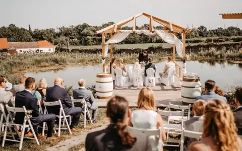 Eine romantische **Hochzeit im Freien** am See. Gäste sitzen auf Stühlen, das Brautpaar steht unter einem Holzdach.