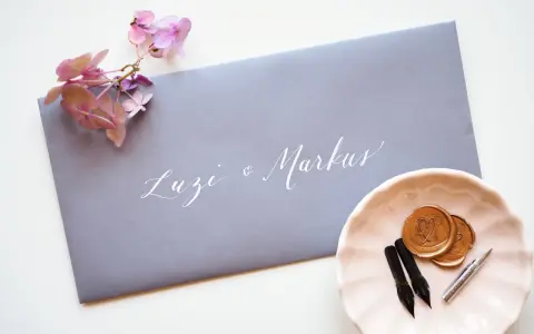 Grauer Umschlag mit weißer Kalligrafie "Luzi & Markus". Daneben eine rosa Schale mit Siegelwachs, Stiften und Federn. Was schreibt man auf den Umschlag zur Hochzeit?