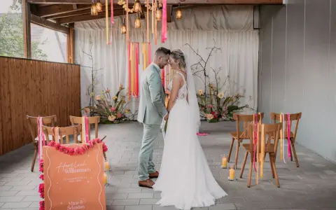 Brautpaar im boho style wedding, umgeben von Blumen und Bändern. Ein Willkommensschild steht im Vordergrund.