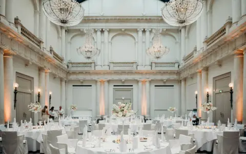 Elegante Tische mit weißen Hussen und Blumenarrangements in einem prunkvollen Saal mit Kronleuchtern – eine besondere Hochzeitslocation.